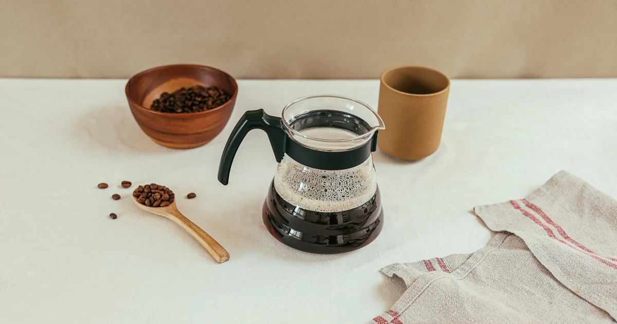 Whole roasted coffee beans beside a glass carafe of freshly brewed drip coffee on a kitchen counter
